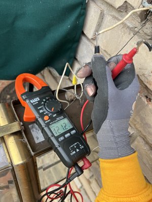 Plugged Electricians ATL — technician using a Klein clamp meter to check live voltage on exterior wiring